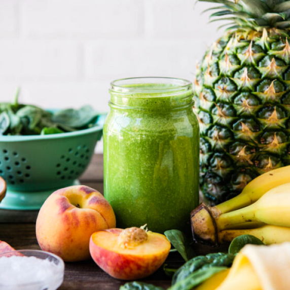 green smoothie in a glass mason jar surronded by bananas, spinach, peaches and a pineapple.