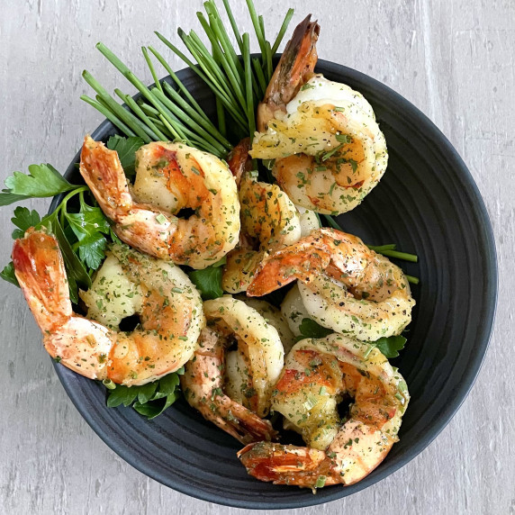 Sautéed shrimps and garnish of fresh herbs in a charcoal bowl on a textured light grey countertop. 