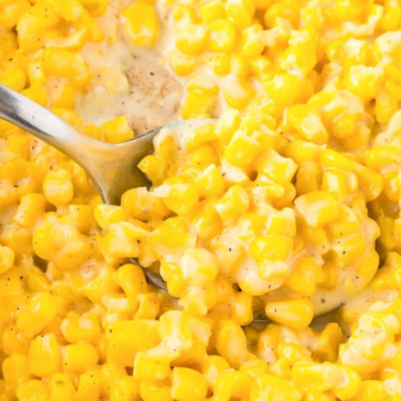 A close-up of a creamy corn dish with yellow kernels with a silver spoon scooping the corn.