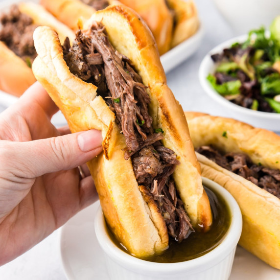 A person is holding a French dip sandwich with beef dipping it into an au jus sauce.