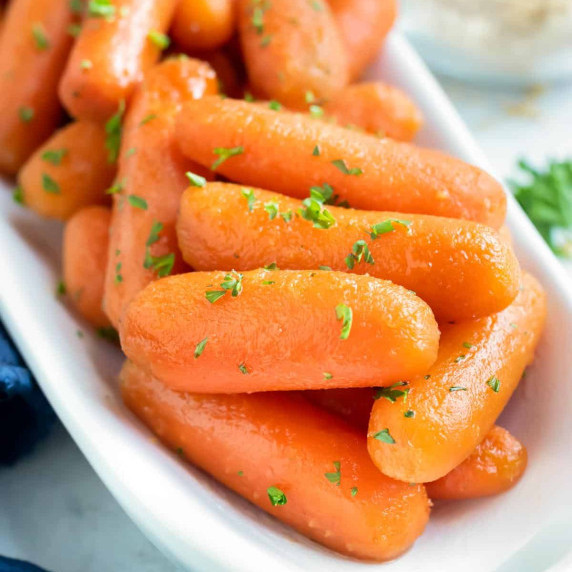 Slow Cooker Glazed Carrots RECIPE in a white serving dish garnished with greens on the top.