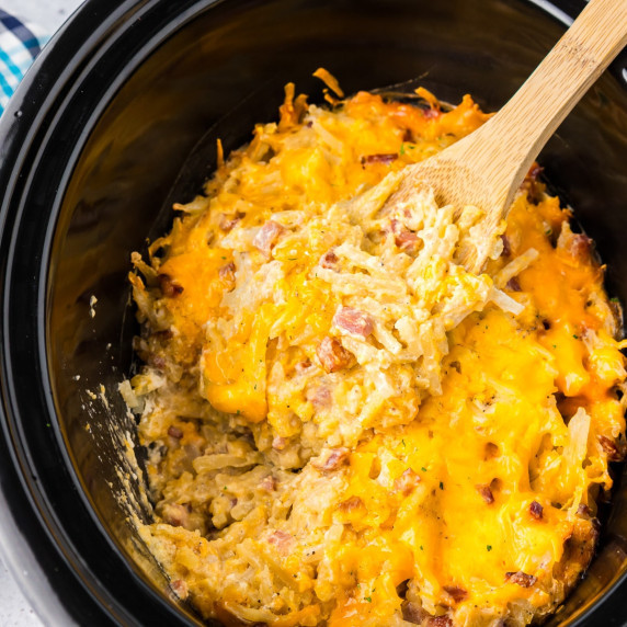 A slow cooker filled with cheesy hashbrown casserole with bits of ham and a wooden spoon.