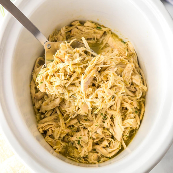 Shredded salsa verde chicken in a slow cooker with a metal serving spoon scooping a spoonful.