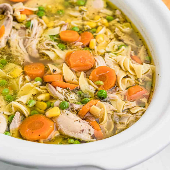 Up close view of an open slow cooker full of chicken, carrrots, peas, corn and noodles in a broth.