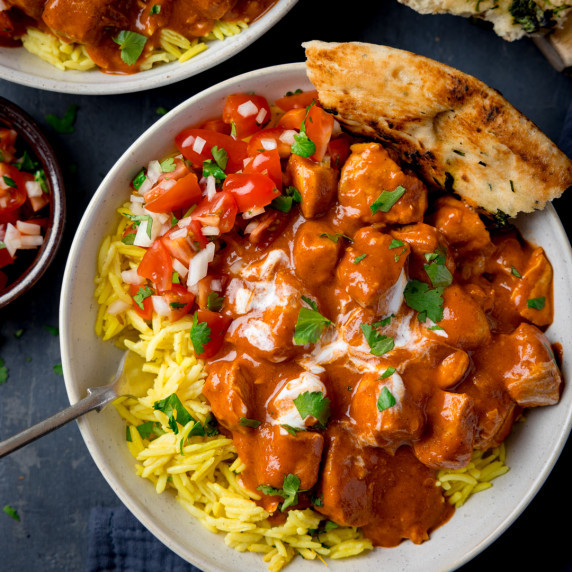 Saucy chicken curry on pilau rice with diced tomato and onion and naan served in a white bowl.