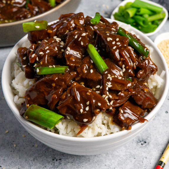 Mogolian beef in a white bowl over rice garnished with sesame seeds and green onions