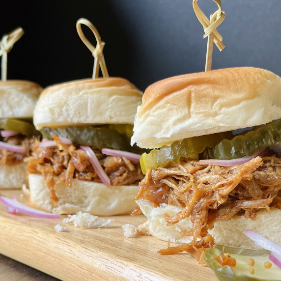 Three pulled chicken slider sandwiches topped with red onion slices and pickles on a wood board.