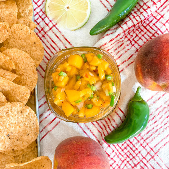 spicy peach salsa on a white/red plaid towel with chips, peaches, jalapeños and lime