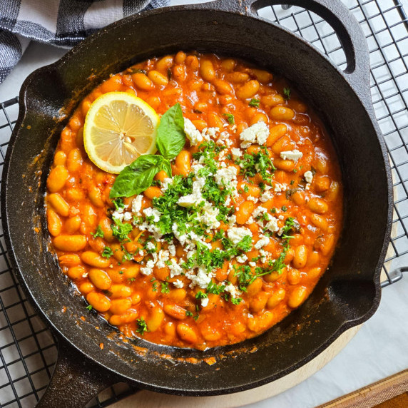 A cast iron skillet full of vibrant, red sauced beans, white feta, and green herbs.