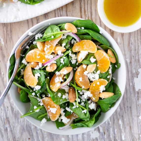 Bowl of spinach salad topped with clementines, almonds and goat cheese with vinaigrette on the side.