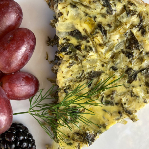 Slice of spinach frittata on a white plate garnished with a dill sprig and served with red grapes.