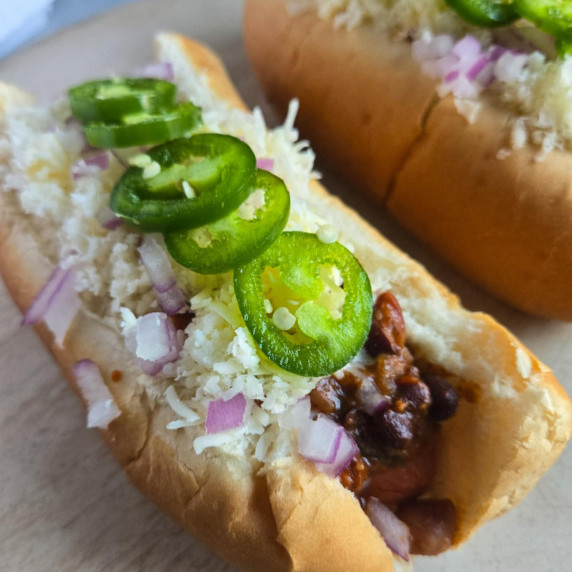 Two buns stuffed with chili, hot dogs, white cheddar, red onions, and green jalapeno slices