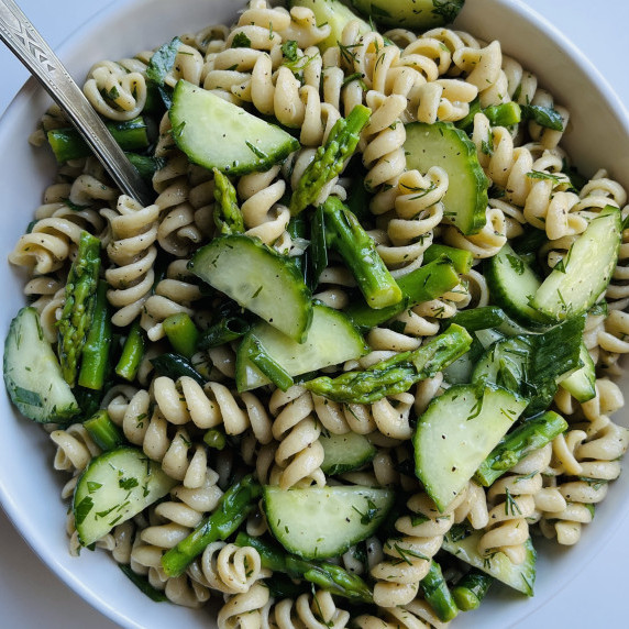 Rotini pasta, cucumbers, green onion and asparagus mixed with an herb dressing in a white bowl.