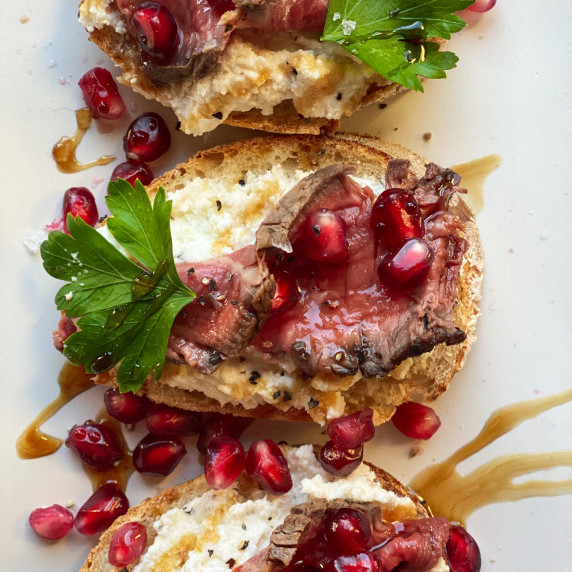 Rare thinly sliced steak on bread with horseradish ricotta, balsamic glaze and pomegranate arils