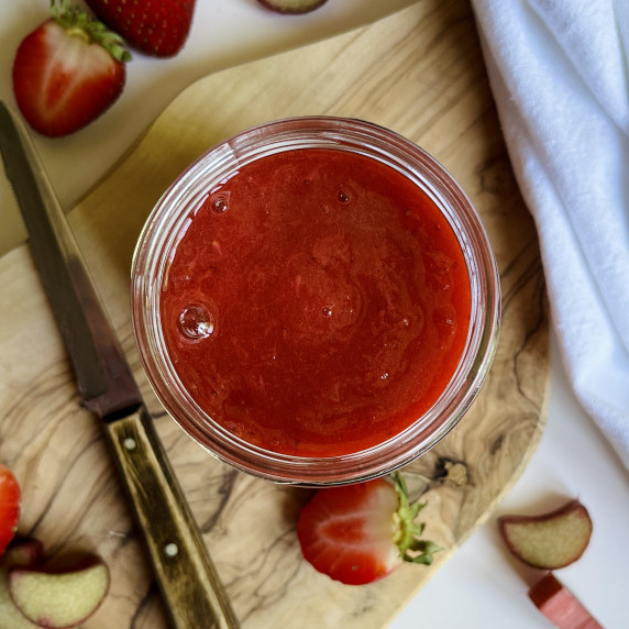 Jar of strawberry rhubarb sauce on a wood cutting with sliced strawberries and rhubarb. 