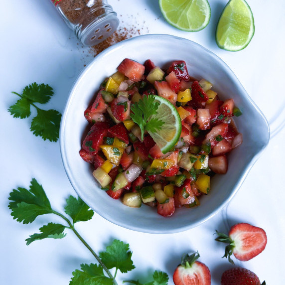 Strawberry salsa in a white bowl surrounded by cilantro and strawberries on a white cutting board.