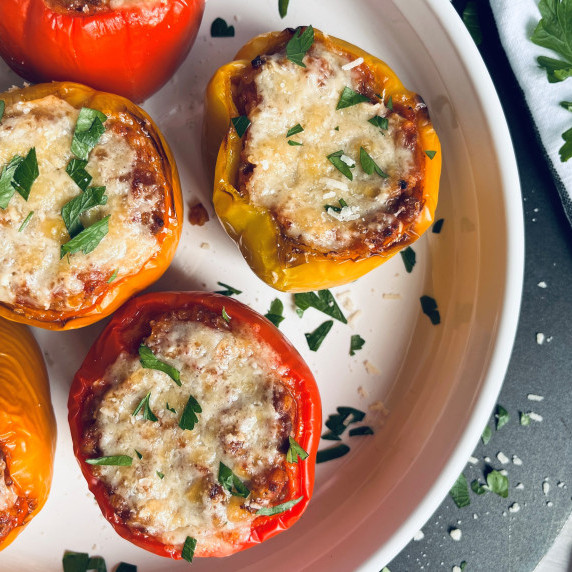 Red and yellow stuffed peppers topped with melted cheese and minced parsley in a white baking dish.