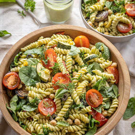 A wooden salad bowl with pasta salad made with fresh vegetables sits on a table scattered with herbs
