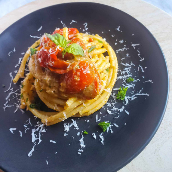 A nest of bucatini garnished with red tomatoes on a black plate against white marble.