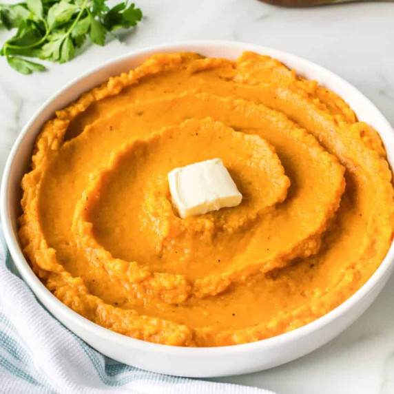 A white bowl filled with mashed sweet potatoes with a square of butter on top.