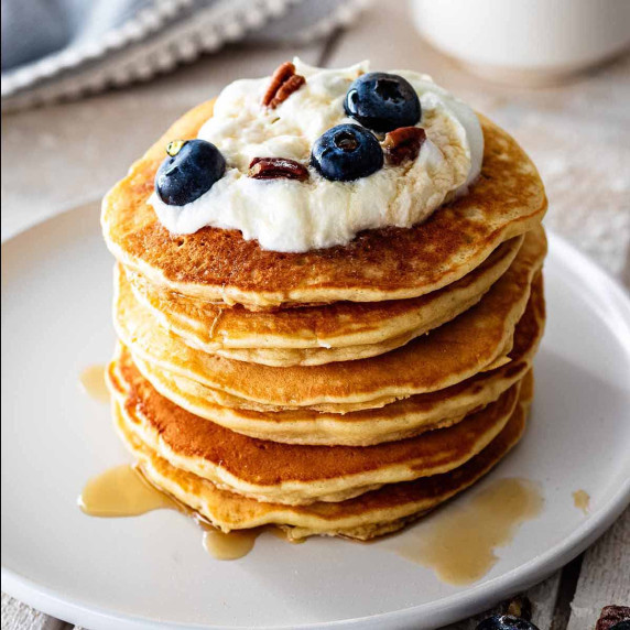 Stack of sweet cream pancakes on a white plate topped with whipped cream, pecans, and blueberries.