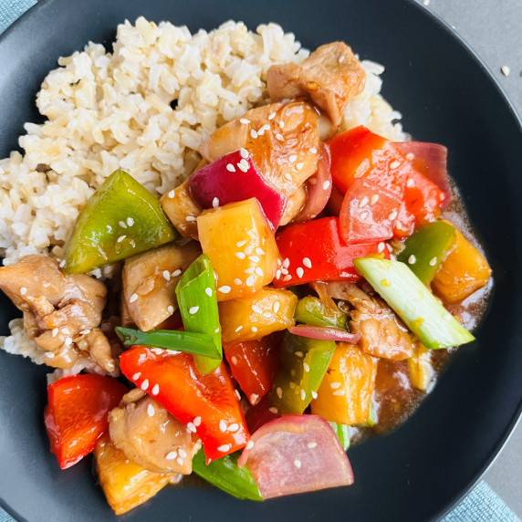 Sweet and sour chicken stir fry with brown rice garnished with sesame seeds on a black plate.