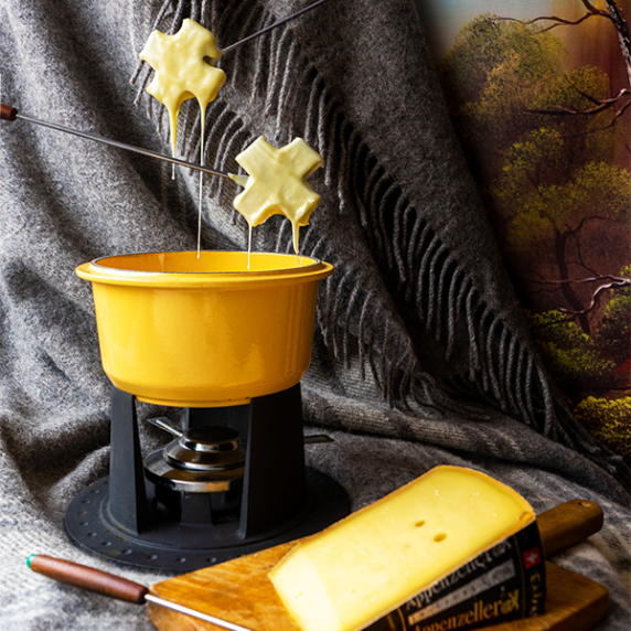 A fondue pot with crosses dripping with cheese hovering over them and a block of cheese in front