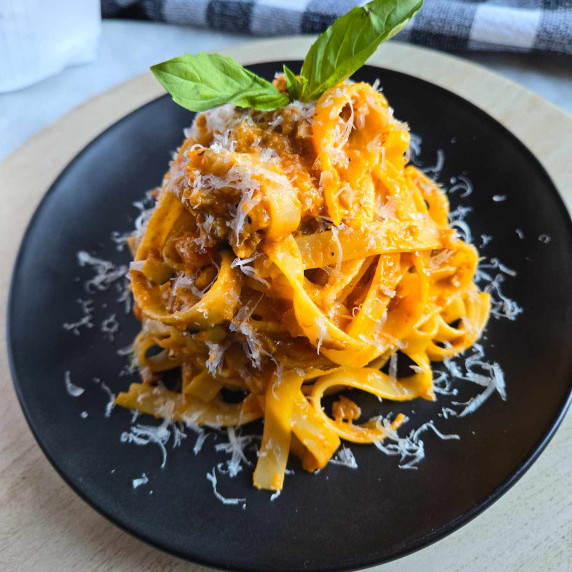 An orangey red hued Tagliatelle Bolognese, topped with green basil leaves on a black plate.