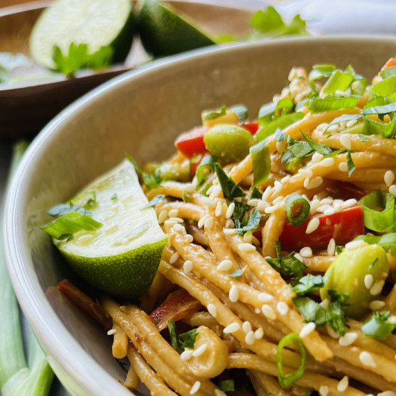 Peanut noodles topped with green onion, sesame seeds, cilantro and a lime wedge in a white bowl.