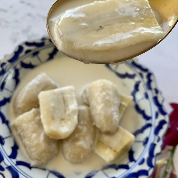A golden spoon with 1 piece of banana above a blue white bowl with bananas in coconut milk sauce.