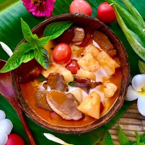 Top-down view of roasted duck curry in a coconut shell with tomatoes and pineapple chunks.