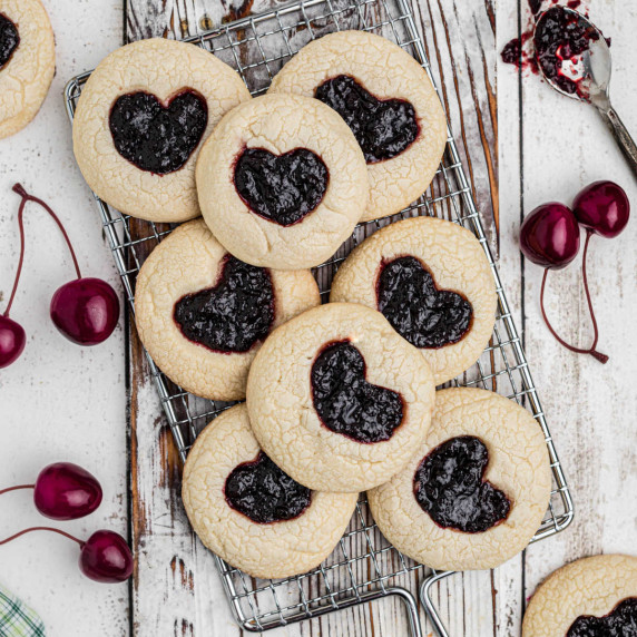 A pile of jam heart cookies on a cooling rack surrounded by cherries.