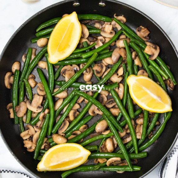 Stir fried green beans and mushrooms on a black pan garnished with lemon slices