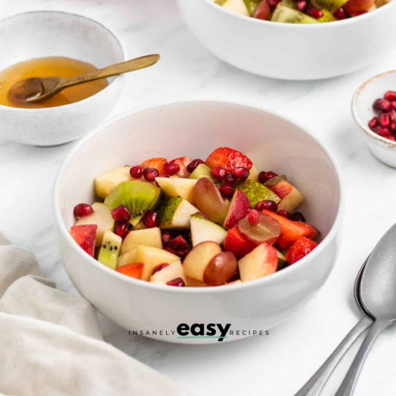 Fruit salad in a white bowl with a bowl of honey at the back and some spoons on the side