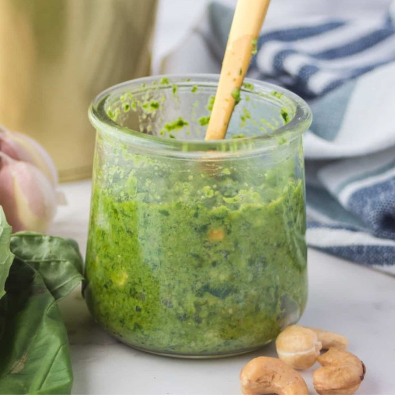 Cashew pesto in a glass container with a wooden spoon, some cashews and basil on the side 