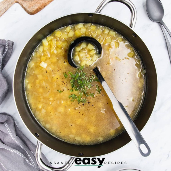 Potato soup topped with fresh chopped parsley and black pepper in a stainless steel pot with a ladle