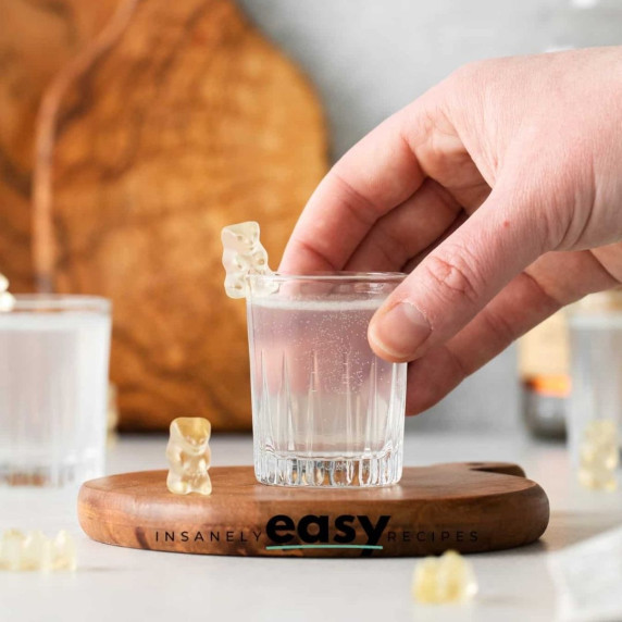 White gummy bear shot in a shot glass served by a hand on a wooden board with a gummy bear beside it