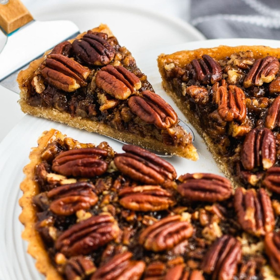 A whole pecan pie on a white plate with one cut slice on a pie server