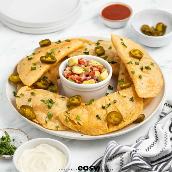 Quesadillas fritas topped with chopped herbs and pickled jalapenos on a plate with avocado salsa