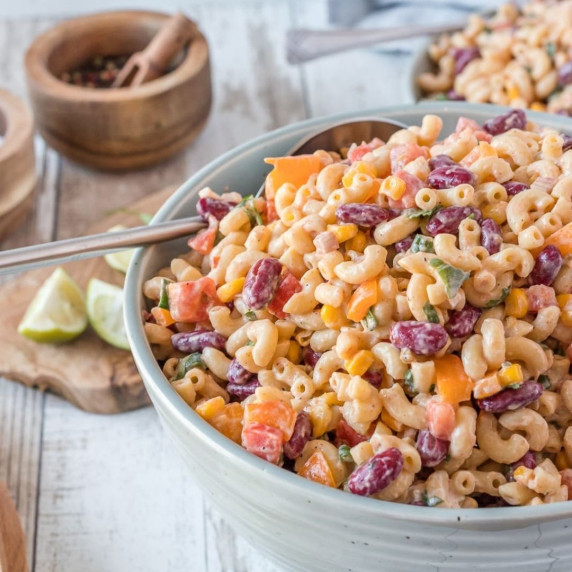 Macaroni salad in a gray bowl with a silver spoon, with lime slices on a wooden board beside