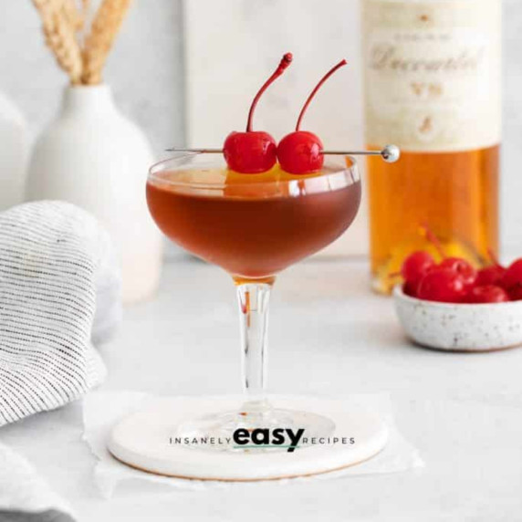 Glass of brandy manhattan cocktail garnished with cherries, with a brandy bottle and cherries behind