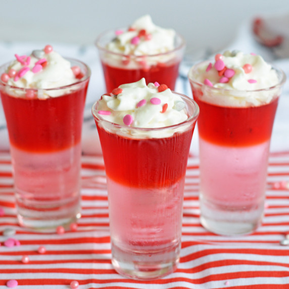 Red and pink layered jello shots with whipped cream and red and pink confetti-style sprinkles.