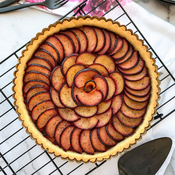 overhead view of a whole vanilla plum tart on a black cooling rack with a white background