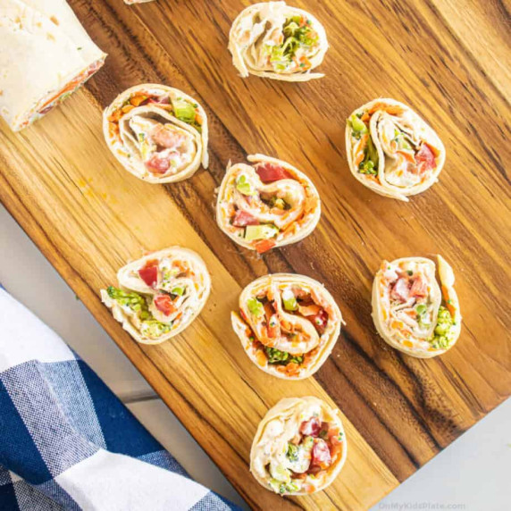 Cutting board from overhead with rolled tortilla pinwheels full of vegetables and a creamy sauce.