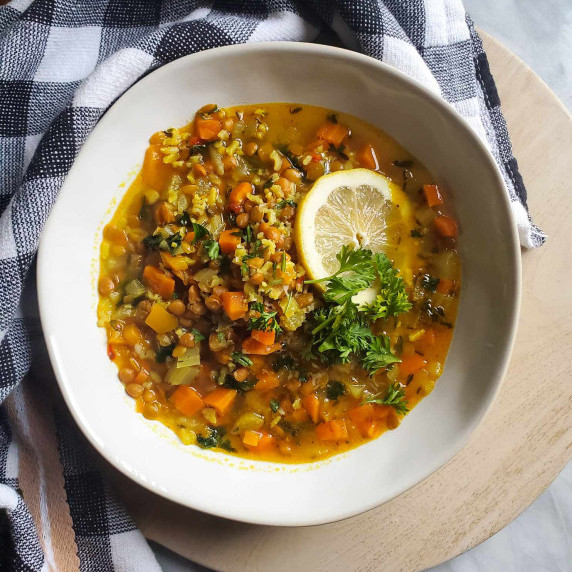 A crisp, white bowl of vegetable lentil soup against a checkered towel on a wooden serving platter