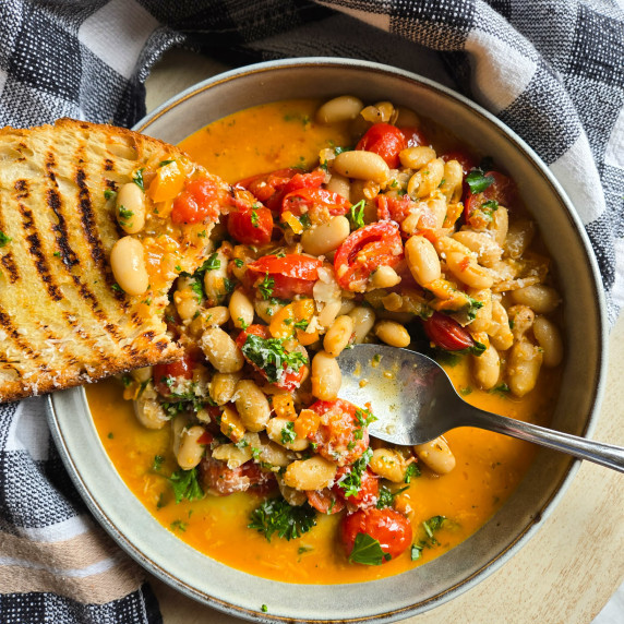 A bowl of brothy beans with pops of green and golden bread against a checkered towel.