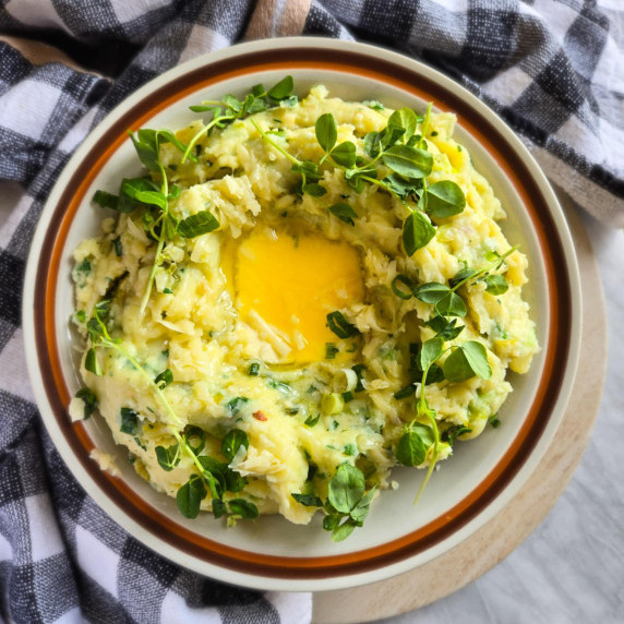 A bowl of fluffy mashed potatoes with pops of green against a checkered towel.