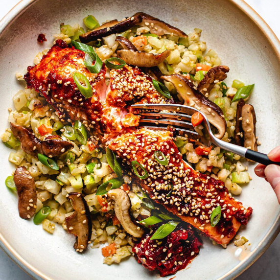 Salmon on a bed of cauliflower rice with scallions, glaze & sesame seeds on a white plate with fork