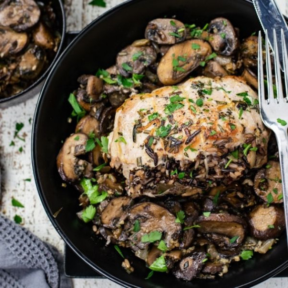 A dish with wild rice stuffed pork chops laying in a bed of mushrooms.