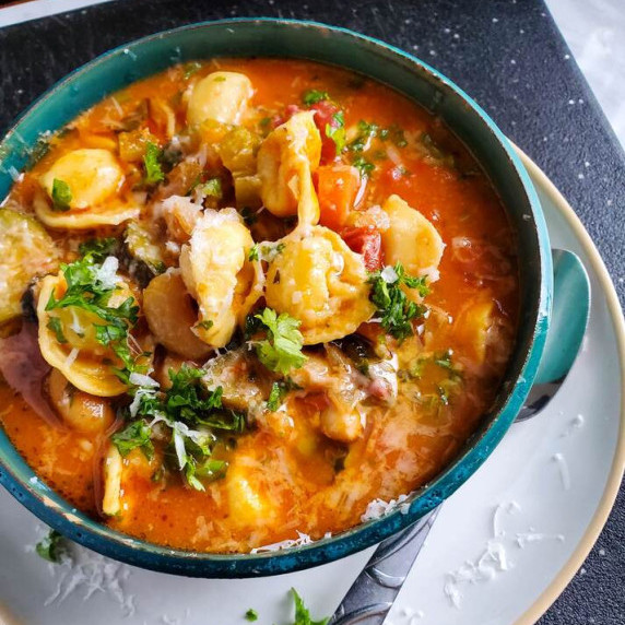 A blue bowl full of red tortellini soup garnished with green herbs and cheese.